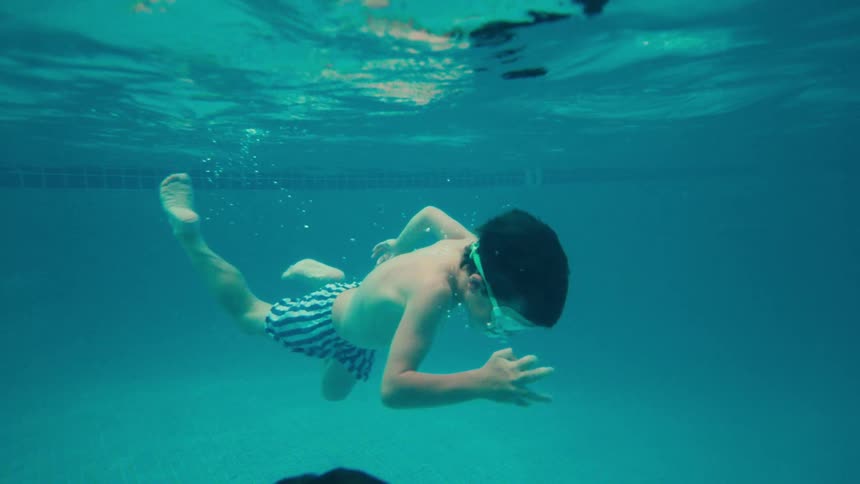 A young boy in goggles gracefully glides through the turquoise water of a swimming pool in slow motion