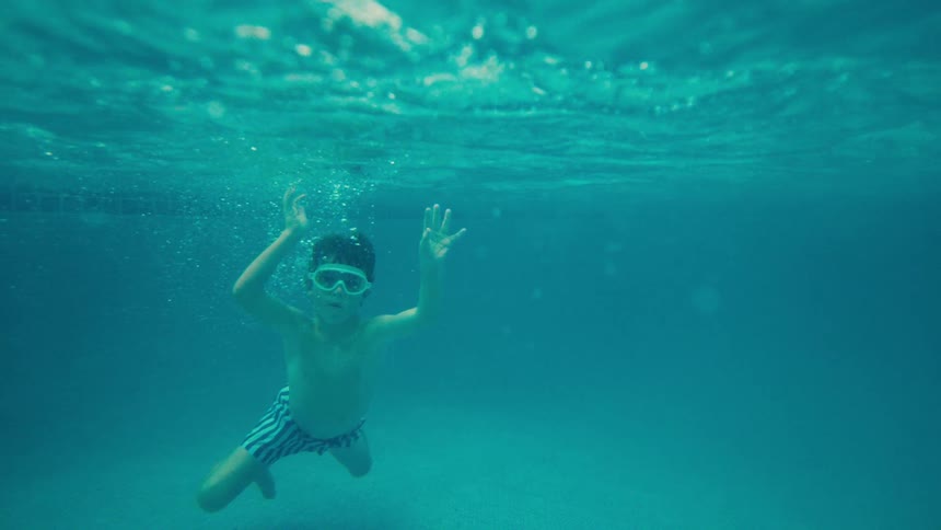 A young boy with swim goggles playfully waves at the camera while floating gracefully underwater in slow motion
