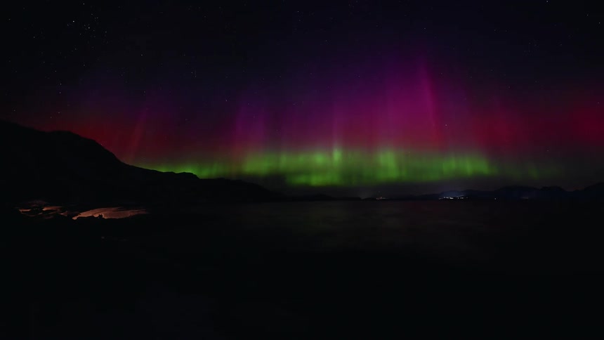 A stunning timelapse of vibrant green and purple Northern Lights dancing over a snowy, mountainous landscape and reflecting in the water below
