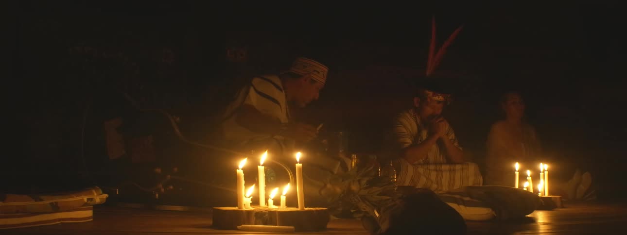 A shaman performs a spiritual healing ritual by candlelight, smoking sacred tobacco and blessing a participant in a dark room