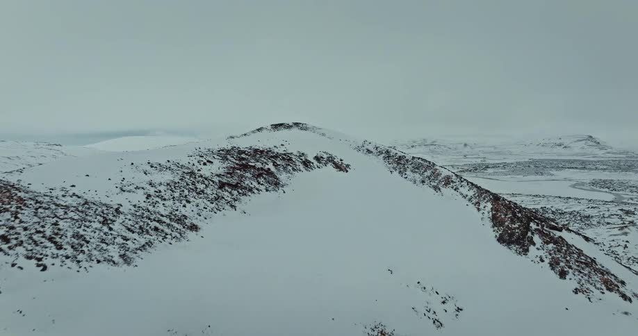 A dramatic aerial reveal shot flies up over a snow-dusted ridge to unveil a vast, frozen volcanic landscape in Iceland