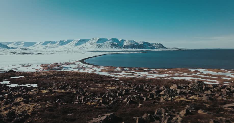An epic aerial view shows a lone traveler standing on a rocky cliff overlooking a vast, snowy bay and mountains in Iceland
