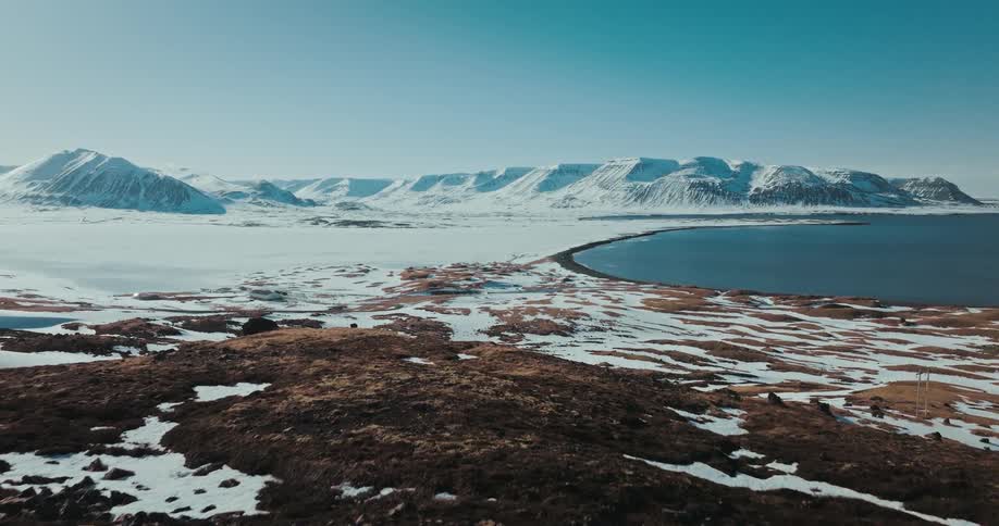An aerial shot flies over a rocky hill to reveal a man standing on the edge, looking out at a vast snowy landscape with a fjord and mountains