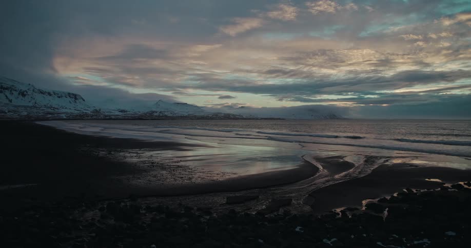 A beautiful wide shot of a tranquil volcanic beach in winter, with snow-covered mountains in the distance under a moody twilight sky