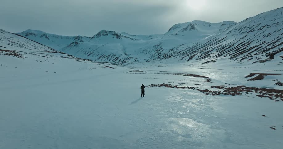 An epic aerial view follows a solitary person walking through a vast, snow-covered valley surrounded by majestic mountains