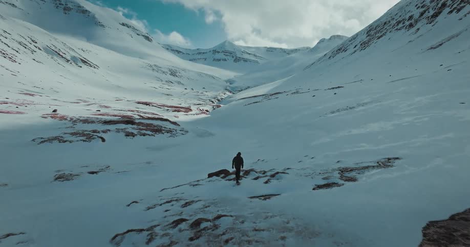 A stunning aerial view tracks a lone adventurer with a snowboard walking through a vast, snowy valley nestled between majestic mountains