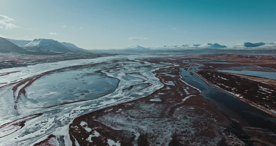 A sweeping aerial view reveals a vast, partially frozen braided river system winding through a valley surrounded by snow-dusted mountains under a clear sky
