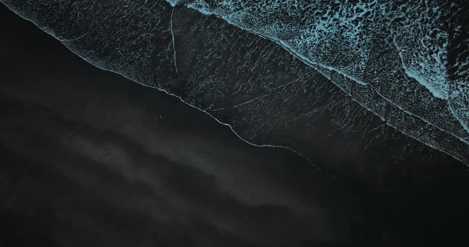 An atmospheric top-down aerial view captures the stark contrast of white sea foam against black volcanic sand as waves gently roll ashore