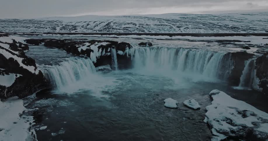 An aerial drone slowly rises to reveal the majestic Godafoss waterfall surrounded by a vast and frozen Icelandic landscape