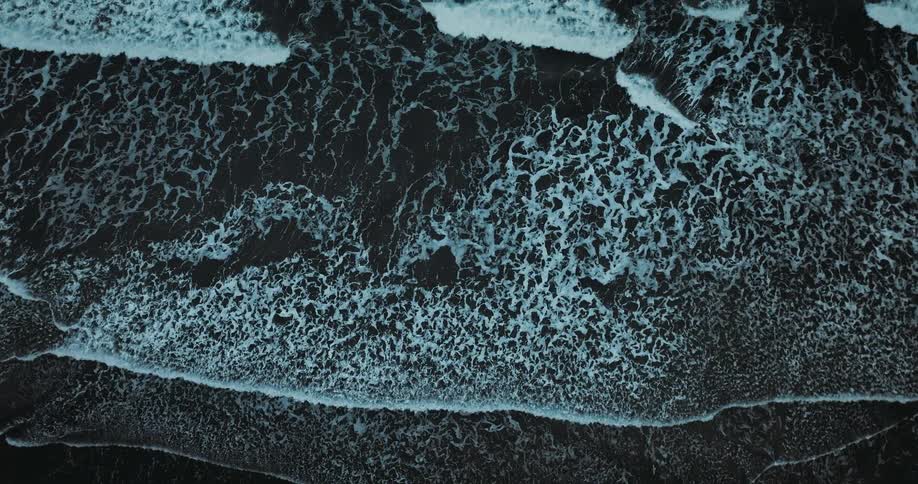 A dramatic top-down slow-motion aerial view of dark ocean waves and white foam washing over a black sand beach