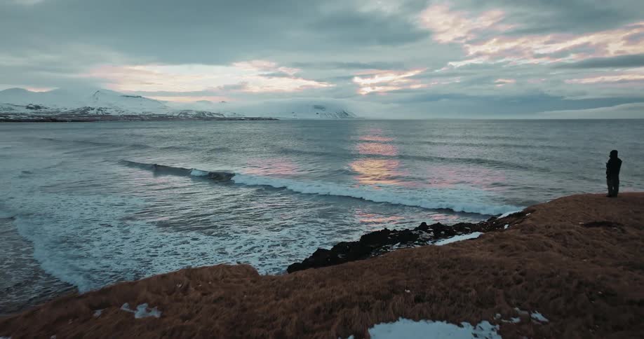 An aerial view glides over a snowy, windswept coastline towards a solitary figure watching the waves crash against the shore at sunset