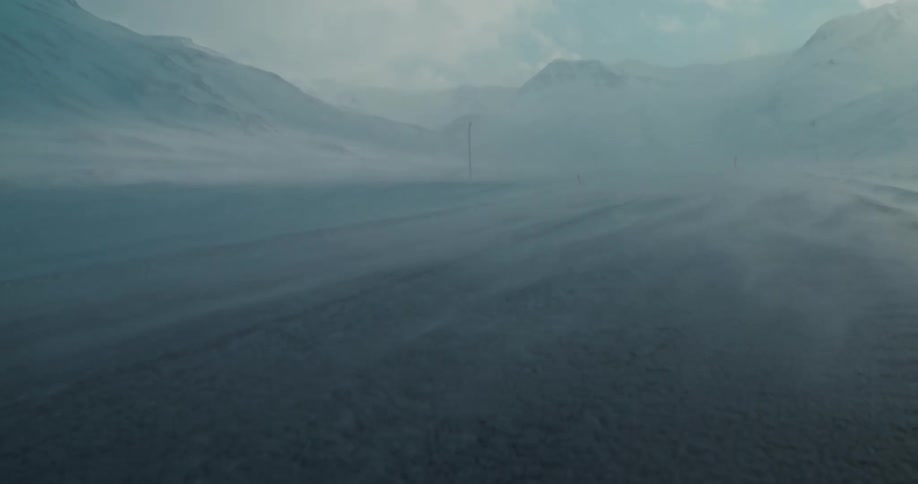 Strong winds blow snow across a frozen, desolate landscape with mountains in the background