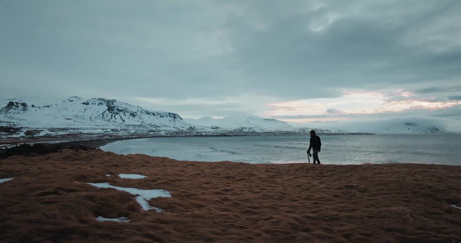 A person with a tripod walks along a snowy, grassy bluff overlooking an icy sea and a snow-covered mountain range at dusk