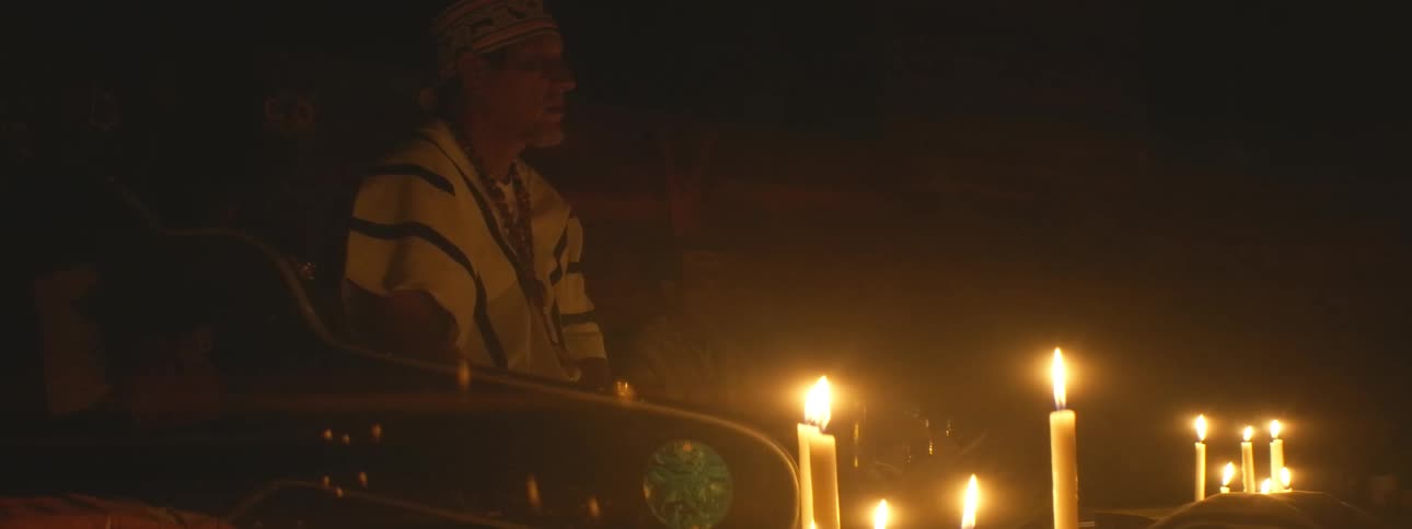 An indigenous shaman in traditional attire performs a spiritual ceremony in a dark room illuminated by flickering candles