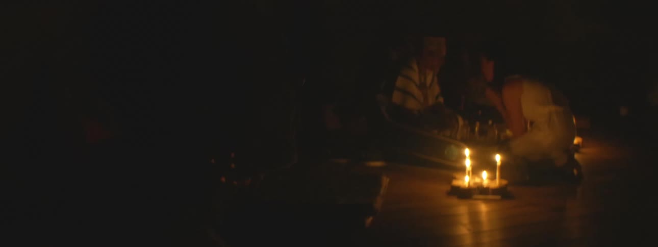 Flickering candlelight illuminates two people sitting in a dark, smoky room, creating an atmospheric and mysterious scene