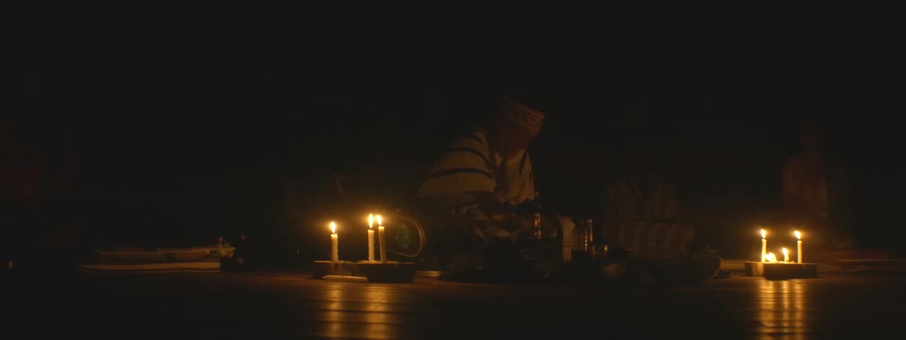 An indigenous shaman in traditional attire performs a spiritual healing ceremony by candlelight in the darkness