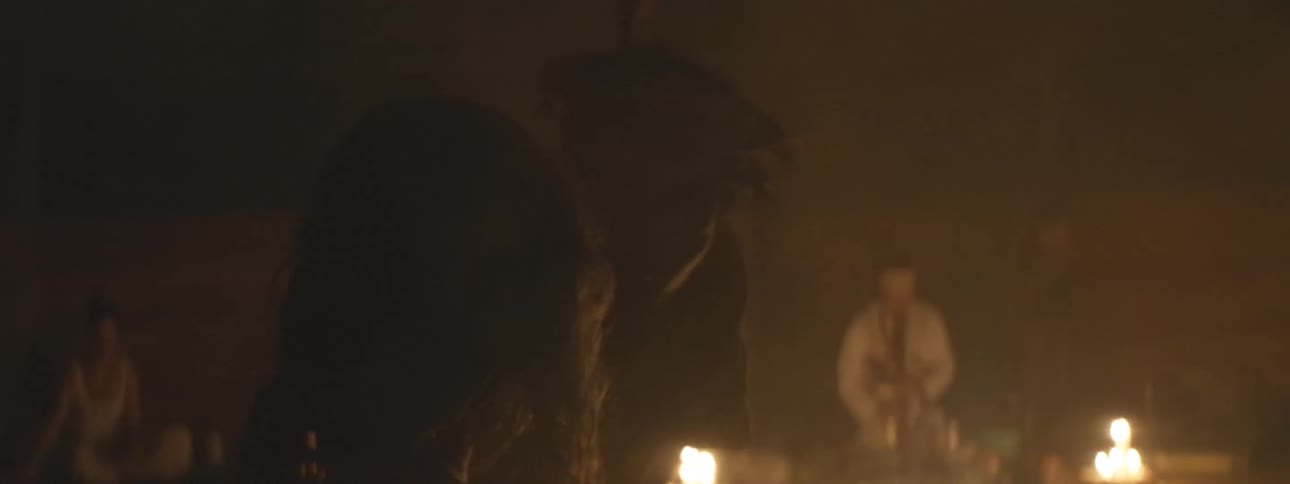 A woman watches as a shaman performs a spiritual ritual in a dark, smoky, candlelit room
