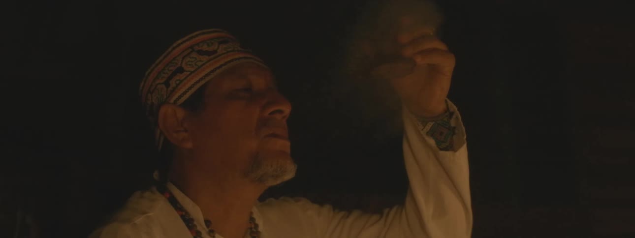 A shaman in traditional attire performs a sacred ritual at night, blowing ceremonial tobacco smoke