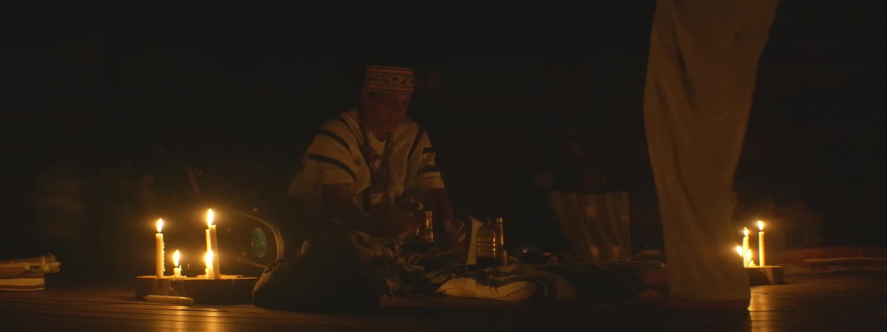 A shaman in traditional clothing performs a spiritual healing ceremony for a person kneeling before him, illuminated by flickering candles in a dark room