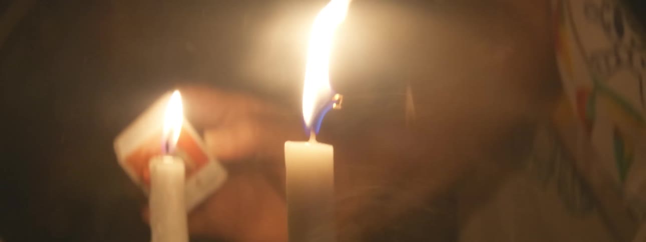 A person blows out two flickering white candles in a dark room, leaving only the glowing wicks before fading to black