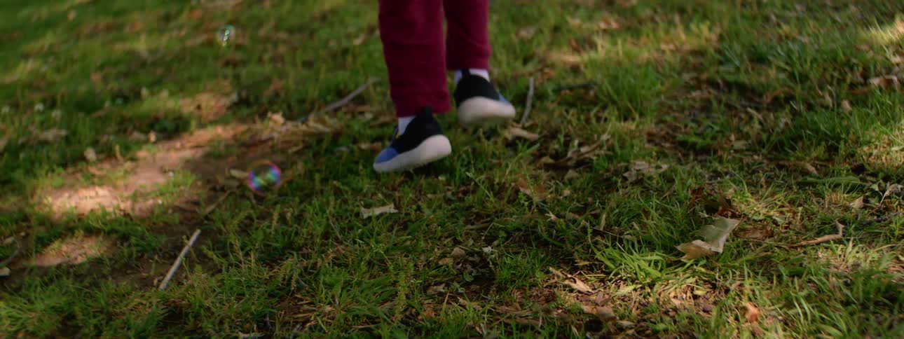 A toddler in a red shirt runs through a grassy park in slow motion, playfully chasing after soap bubbles
