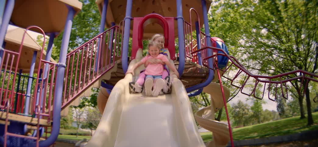 Two happy young sisters smile as they go down a colorful playground slide together in slow motion on a sunny day