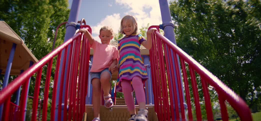 A low-angle, slow-motion shot of two adorable little girls holding hands and smiling as they descend the stairs at a colorful park playground