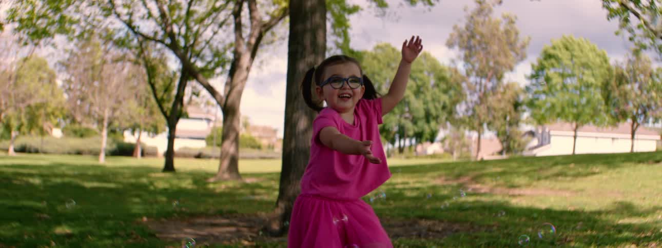 A joyous little girl in a bright pink outfit and glasses chases soap bubbles in a park in slow motion