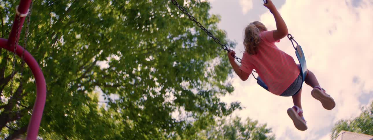 Low-angle slow-motion shot of a young girl with blonde hair enjoying a swing set on a sunny summer day