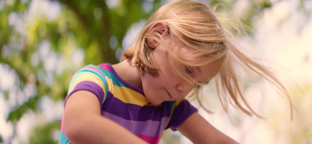 A close-up shot captures a young blonde girl's hair blowing gently in the wind while she is focused on playing outside