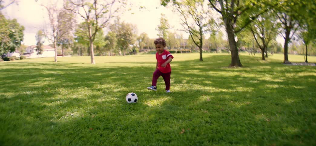 An adorable toddler joyfully runs and plays with a soccer ball in a sun-drenched park in slow motion