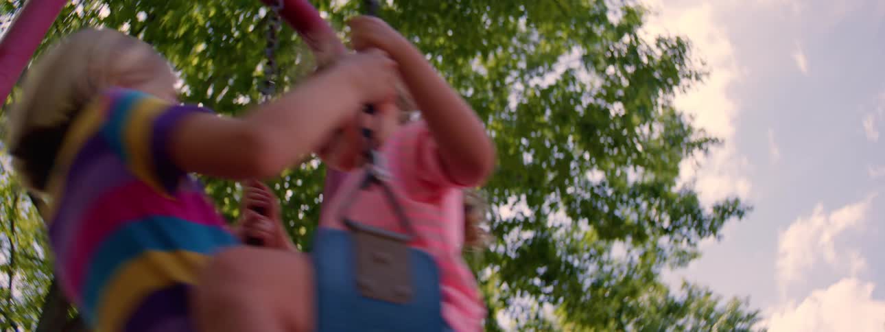 Two young girls smile and laugh as they swing back and forth in slow motion on a playground swing set