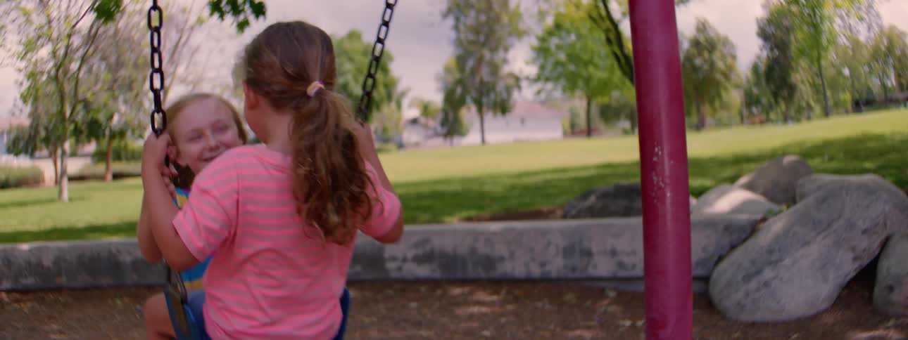 Two happy young girls smile and swing together at a park in a nostalgic slow-motion shot