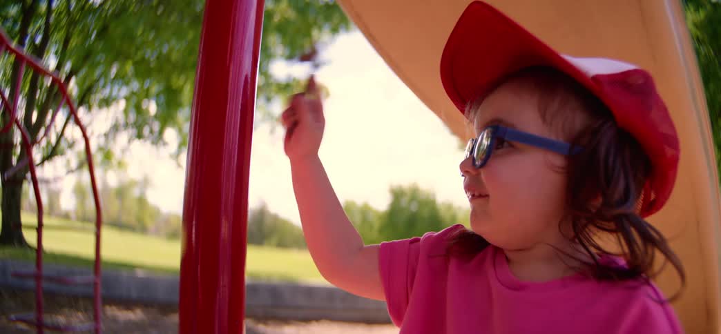 Slow motion close-up of a happy little girl in a red hat and glasses playing with a stick at a sunny park playground