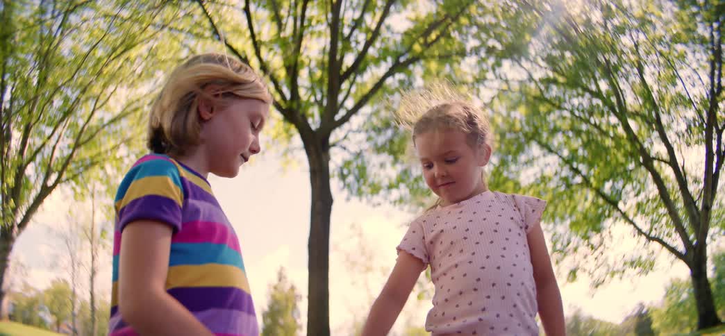 A fisheye, slow-motion shot from a low angle shows two young sisters holding hands and walking together in a sunny park