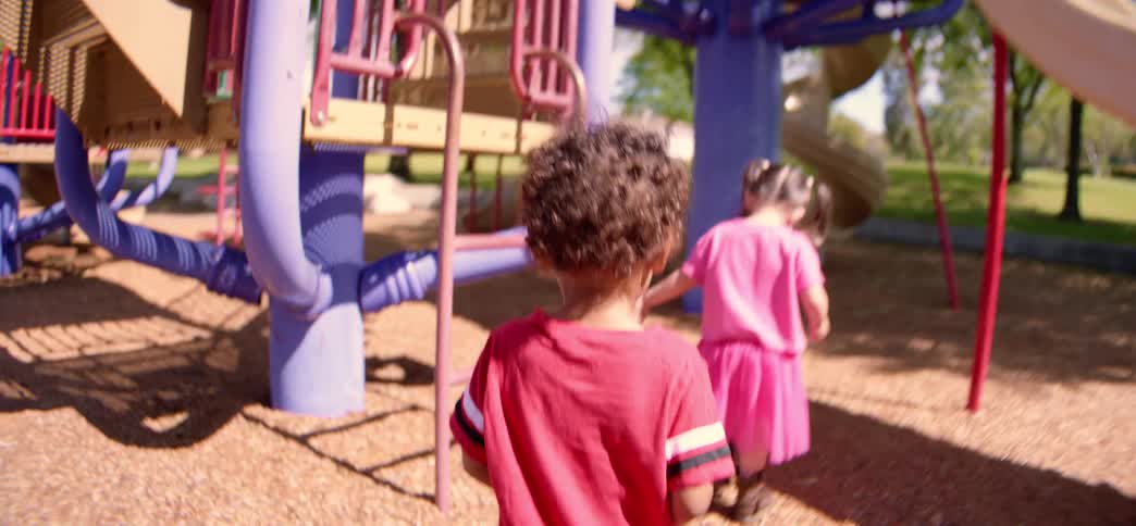 A young boy follows a little girl in a pink dress as they walk across a sunny playground in slow motion