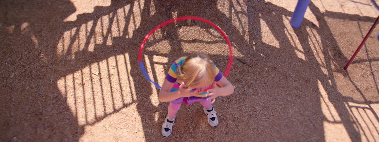 A young girl in a brightly colored dress tries to hula hoop at a playground in slow motion, shot from a high angle