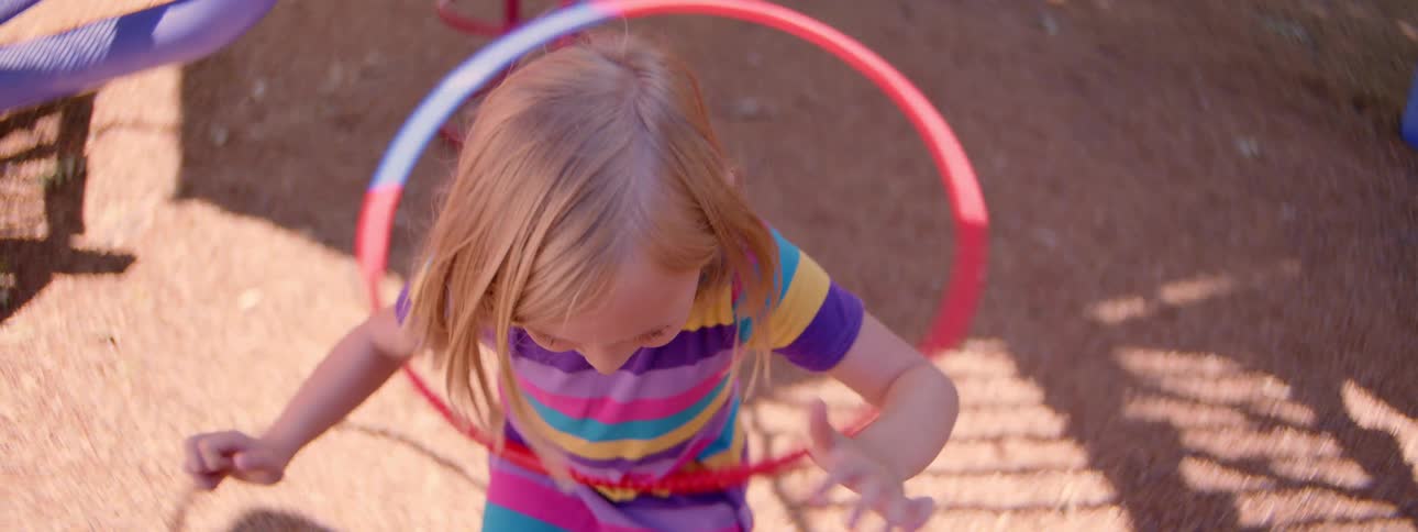 High-angle, slow-motion footage shows a little blonde girl in a brightly striped shirt spinning a hula hoop at a sunny playground