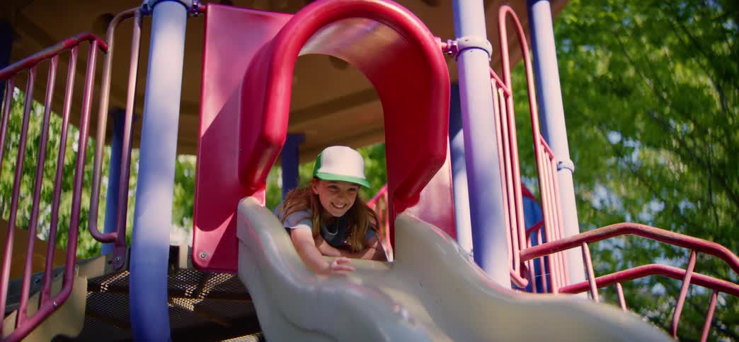 A happy young girl wearing a trucker hat smiles as she prepares to slide down a colorful playground slide