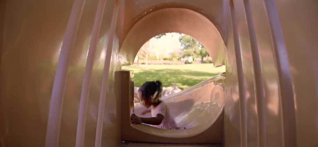 A playful African American toddler waves goodbye from inside a playground tunnel slide at a park on a sunny day