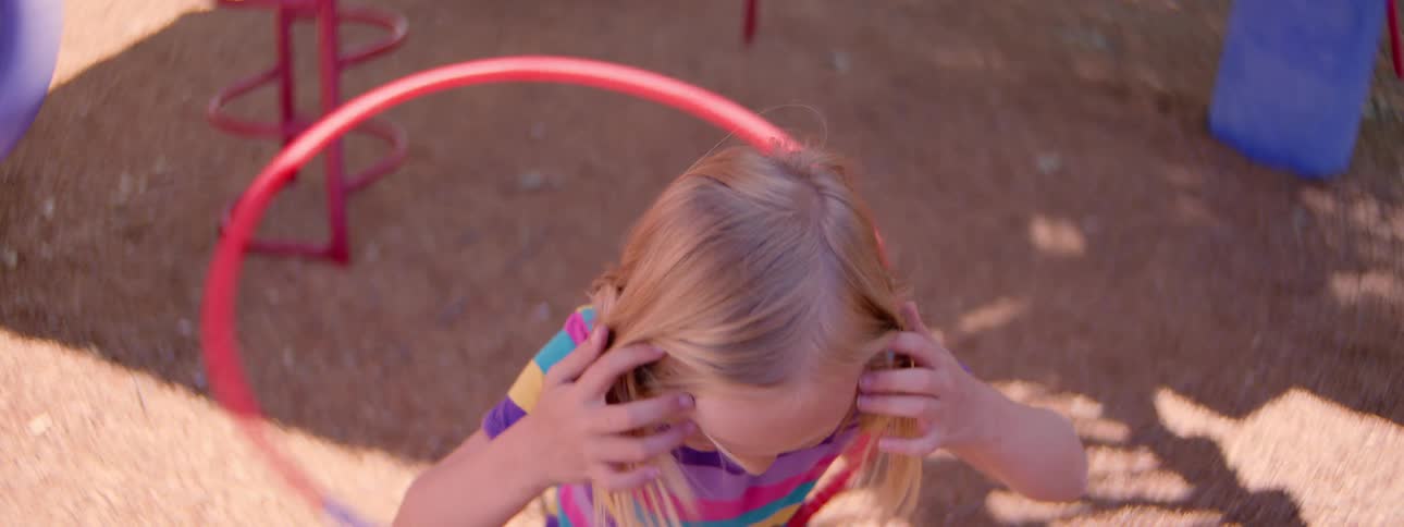 A young girl in a colorful striped shirt plays with a hula hoop in slow motion from an overhead perspective on a playground