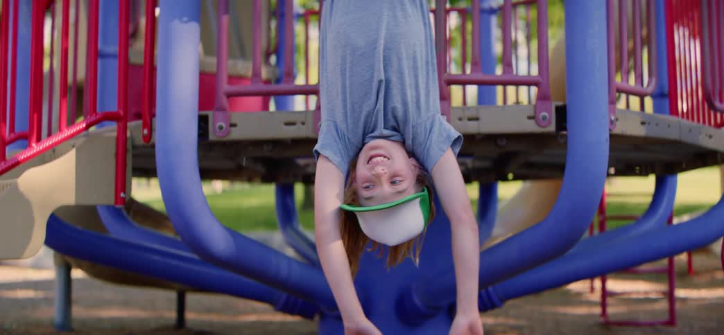 A playful boy in a trucker hat hangs upside down from a colorful jungle gym and smiles happily for the camera