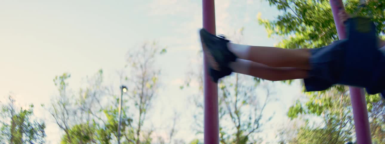 Slow motion low angle shot of a young boy in a green hat swinging high into the sky at the playground