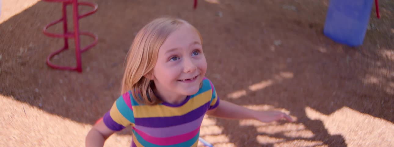 A happy young girl with a missing tooth smiles and waves while a hula hoop spins around her in slow motion at the playground