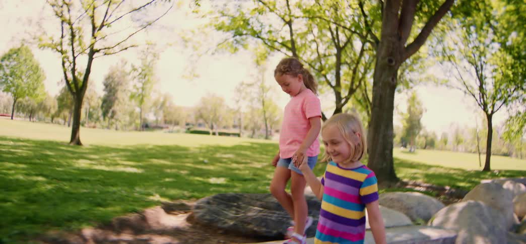 A young girl in a pink shirt carefully balances on park rocks until her friend joins her, and they walk hand-in-hand along a low wall