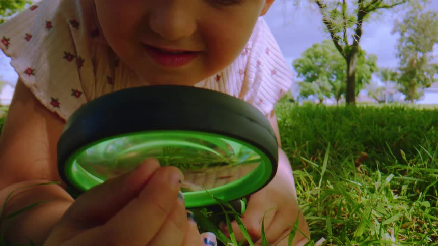 A curious little girl with blonde curly hair lies in the green grass, discovering the wonders of nature through a magnifying glass