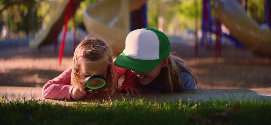 Two curious young girls lie on their stomachs at a sunny park, using a magnifying glass to explore tiny plants and bugs in the grass