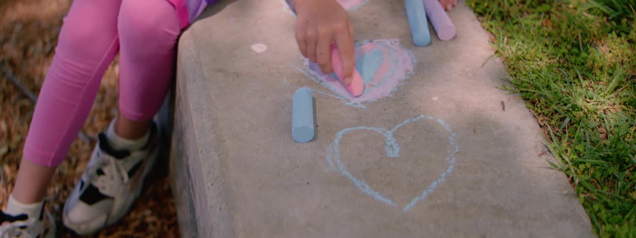 A close-up shot of a young child's hand coloring in a vibrant pink and blue heart with chalk on a concrete sidewalk