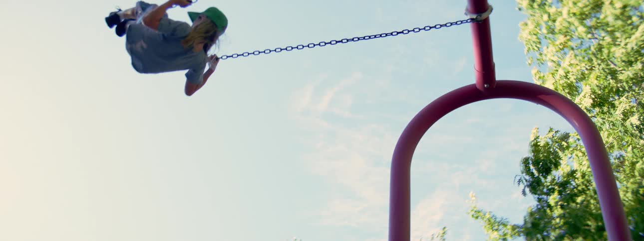 A slow-motion, low-angle shot of a young boy in a baseball cap swinging high into the bright blue sky