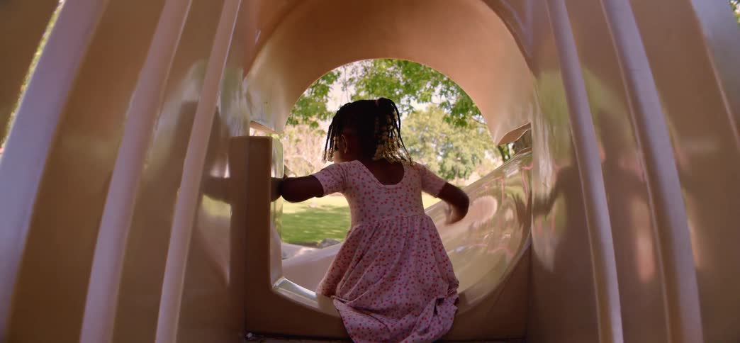 A joyful little girl crawls through a playground tunnel, turns to smile at the camera, and then slides down the slide in slow motion